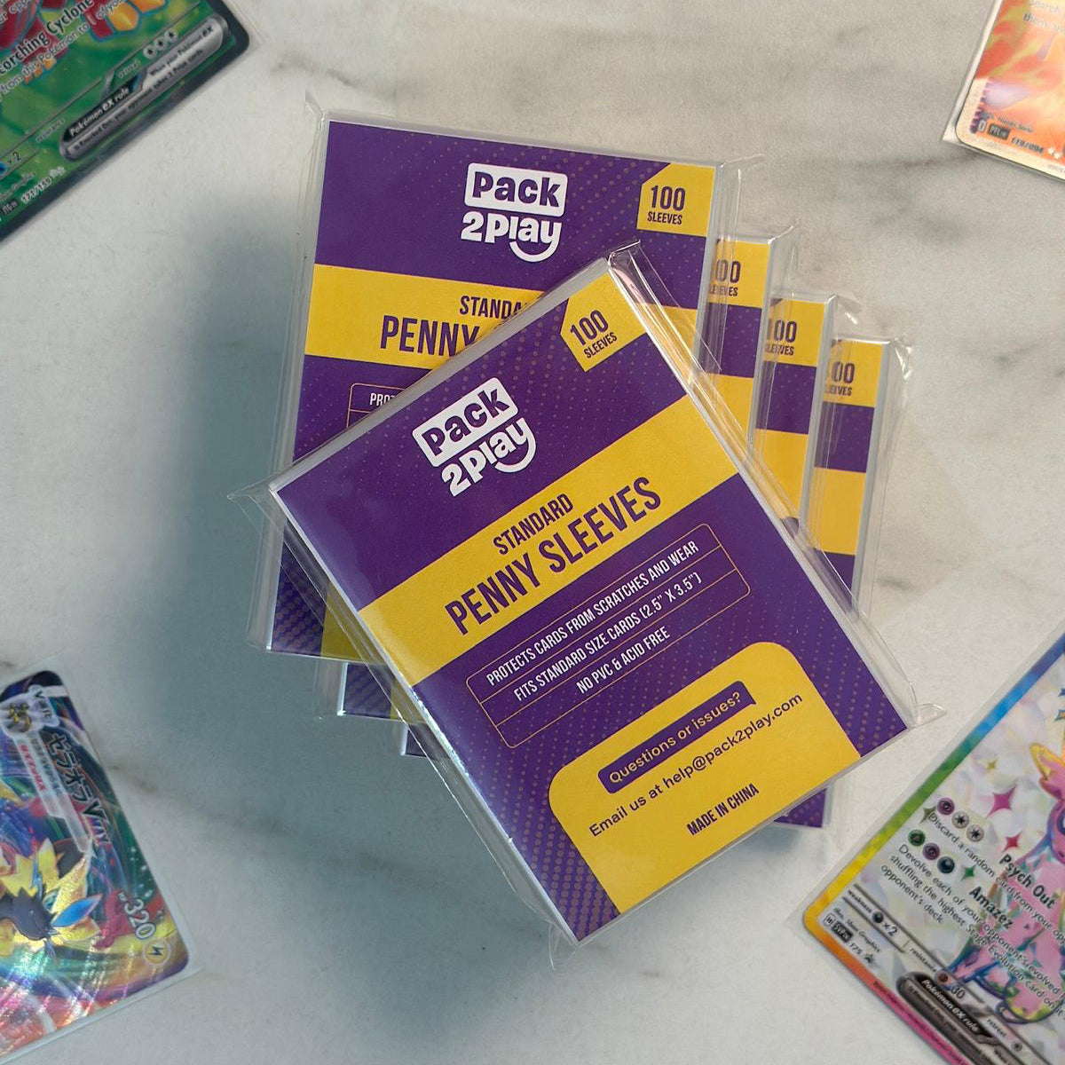 Two packs of Pack2Play Standard Penny Sleeves on a marble surface with trading cards around.